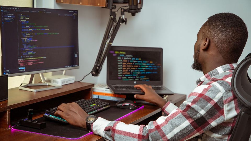 A software developer in Nairobi writing code across a laptop and a desktop monitor